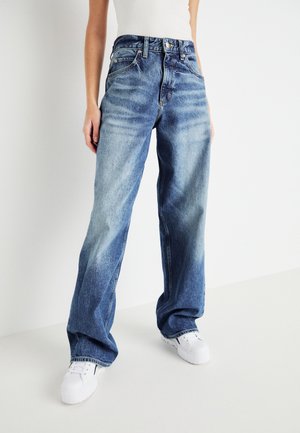 Person wearing loose-fit, faded blue jeans and white sneakers, standing against a plain white background.