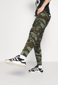 Vihreät ja ruskeat maastokuvioidut cargo-housut, joissa sivutaskut, joustavat lahkeensuut ja kapeneva leikkaus, yhdistetty mustavalkoisiin lenkkareihin.