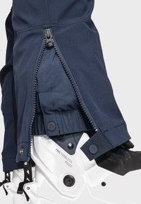 Navy wasserdichte Jacke im Nahaufnahme mit seitlichem Reißverschluss, elastischen Bündchen und Details über einem weißen Stiefel mit reibungsfreier Drehhardware.