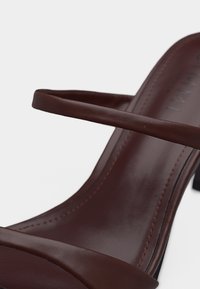 Καφέ δερμάτινο πέδιλο με ψηλό τακούνι, λεπτά λουριά και εμφανή ραφές κατά μήκος της σόλας, σε μονόχρωμο φόντο.