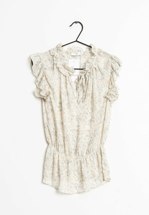 Blouse florale couleur crème avec manches et encolure volantées, taille cintrée, suspendue à un cintre noir sur fond blanc.