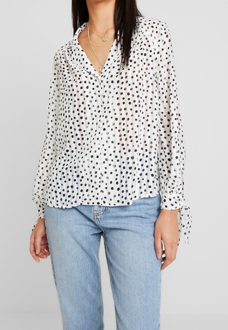 Blusa trasparente bianca con pois neri, chiusura a bottoni sul davanti, maniche lunghe con dettagli a fiocco e vestibilità comoda. Abbinata a jeans azzurri chiari.