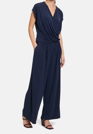 Kvinde iført en mørkeblå ærmeløs jumpsuit med draperet V-hals og vidde bukseben, sat sammen med sorte åbne kilehælssandaler.