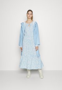 Capa de chuva azul claro com capuz sobre um vestido maxi floral em tons semelhantes. Tecido suave, bainha com babados, e combinada com botas de borracha brancas.