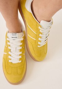 Sneakers in suede giallo con accenti bianchi. Punta arrotondata, design con lacci e suola in gomma color beige. L'etichetta del marchio è visibile sulla linguetta.