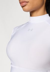 Λευκό αθλητικό τοπ με ψηλό λαιμό και κοντά μανίκια, με διακριτικό γκρι ανοιχτό λογότυπο στο στήθος και ομαλή, εφαρμοστή υφή.