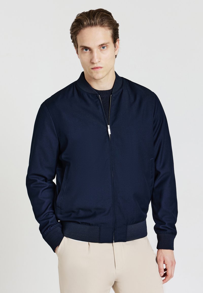 Jeune homme à la peau claire et aux cheveux peignés en arrière, portant une veste bomber navy sur un pull sombre et un pantalon beige, se tenant avec une main dans la poche.