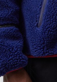 Fleece pullover i mørkeblå med høj krave, lynlås foran og kontrasterende rød kant. Har en blød tekstur og sorte syninger.