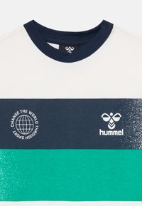 Fehér póló tengerészkék gallérral, amely tengerészkék és türkiz vízszintes csíkokat, valamint egy földgömböt ábrázoló logókat tartalmaz "Change the World Through Sport" felirattal.