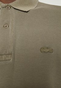 Khaki polo shirt with a textured fabric, featuring three buttons at the collar and a green embroidered crocodile logo on the chest.