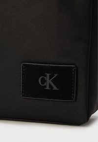 Μαύρη τσάντα από ύφασμα με κομψό σχεδιασμό και υφή δερμάτινου μαύρου λογότυπου με ανάγλυφο το σήμα «cK». Μινιμαλιστική και σύγχρονη.