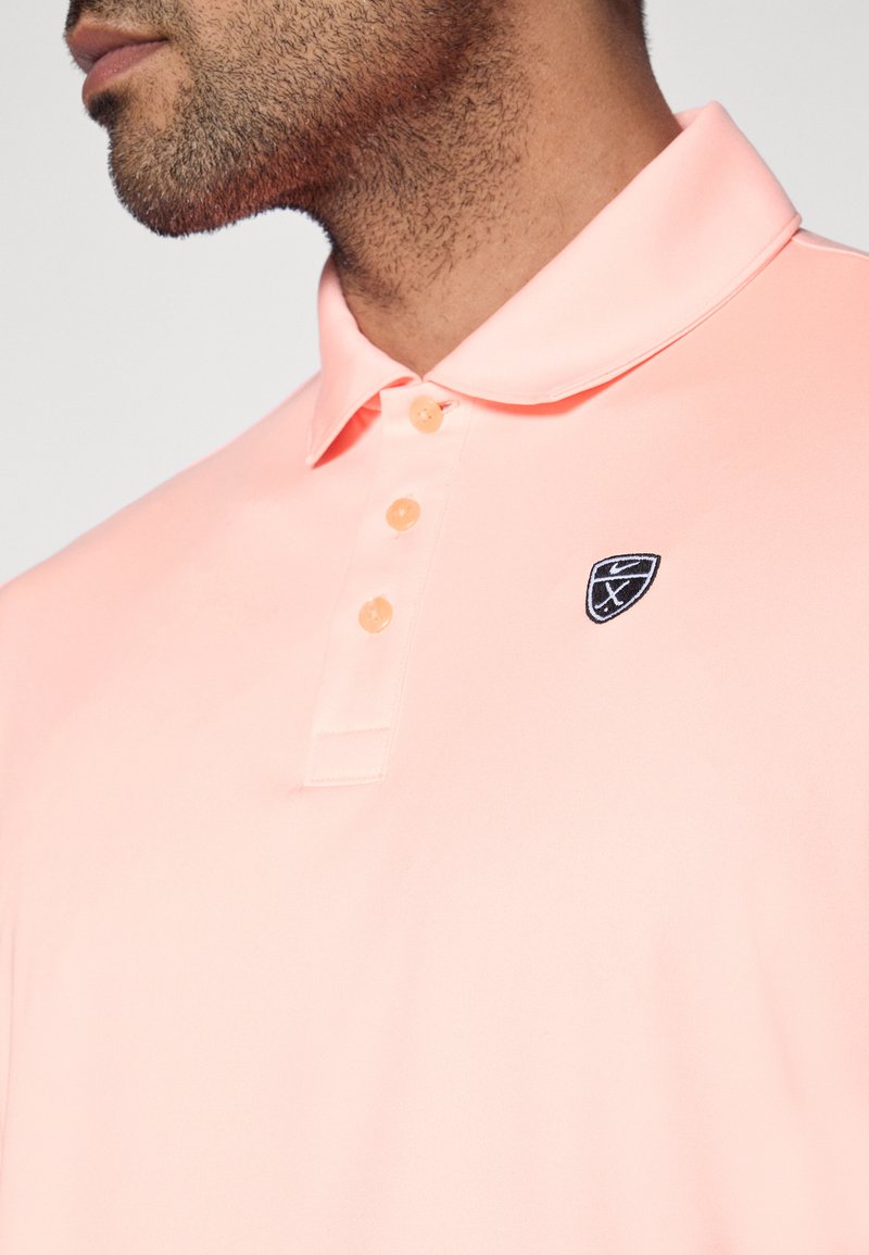 Polo à manches courtes de couleur rose clair, avec une patte de boutonnage à trois boutons et un patch logo noir sur le côté gauche de la poitrine. Texture de tissu lisse.