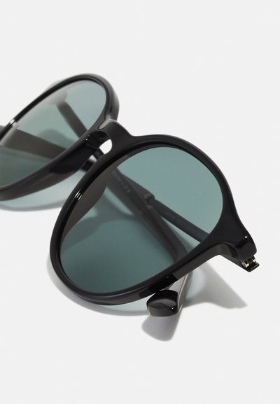 Mustat aurinkolasit pyöreillä kehillä, liukuväri vihreillä linsseillä ja sulavalinjaisilla varsilla. Ominaisuuksiin kuuluu minimalistinen muotoilu ja korostetut saranat.