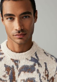 Knit sweater with a cream base, featuring a multicolored abstract pattern in brown and blue, round ribbed neckline, and soft texture.