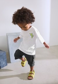 T-shirt blanc à manches courtes avec un motif de palmier vert, associé à un short gris foncé et des sandales jaunes à deux boucles.