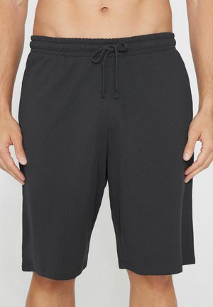 Schwarze knielange Shorts mit elastischem Bund und Kordelzug, getragen von einer stehenden Person mit entspannten Händen an den Seiten.