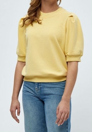 Osoba ubrana w żółtą bluzkę z krótkimi rękawami i bufkami na ramionach, w połączeniu z niebieskimi dżinsami z wysokim stanem, stojąca na jednolitym tle.