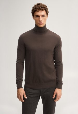 Mann mit lockigem braunem Haar, der einen eng anliegenden dunkelbraunen Rollkragenpullover und dunkle Hosen trägt, stehend vor einem schlichten hellen Hintergrund.