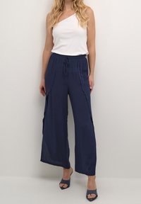 Marineblauwe wijde broek van lichtgewicht stof, met een trekkoord in de taille en zijsplitten. G gedragen met een witte top met één schouder.