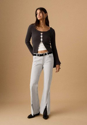 Frau trägt eine schwarze, geknöpfte Strickjacke über einem weißen Crop-Top, hellgraue Jeans mit geteiltem Saum, schwarzen Gürtel und schwarze Stiefel, steht vor einem beigen Hintergrund.