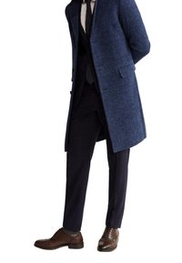 Uomo che indossa un soprabito blu, pantaloni scuri, camicia bianca, cravatta nera e scarpe di pelle marroni, in piedi con le mani dietro la schiena.