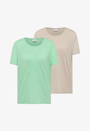 Femme souriante aux longs cheveux bruns, portant une chemise à manches courtes vert clair et tenant une veste beige dans sa main gauche.