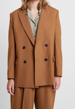 Žena v hnědém plášti s dvojitým knoflíkováním a černými knoflíky přes světle zelenou košili na knoflíky a odpovídající hnědé kalhoty.