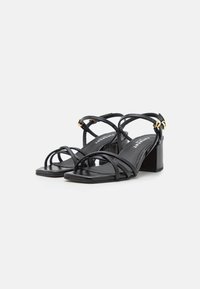 Sandales noires à lanières fines en cuir, avec un bout carré et un talon moyen bloc. Des détails en métal doré viennent sublimer le design.