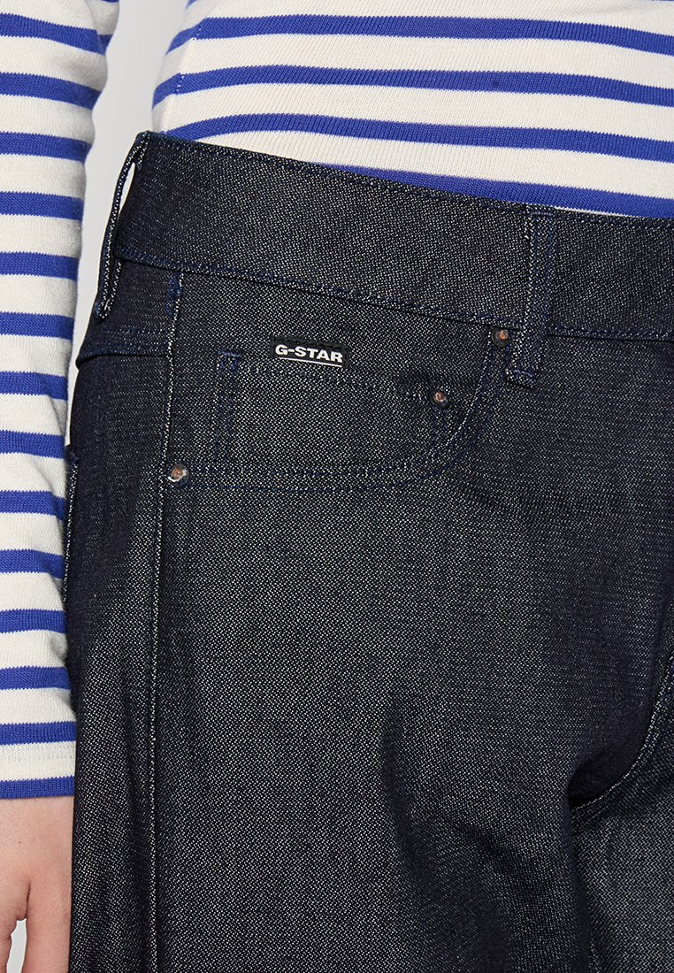 Jean en denim bleu foncé avec une finition texturée, arborant une étiquette G-Star, des poches avant et des accessoires en métal. Porté avec une chemise à rayures à manches longues.