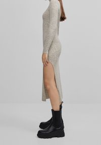Robe longue en tricot beige à manches longues avec fente latérale, associée à des bottines noires épaisses avec semelle texturée et languette.
