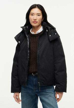 Kvinde iført en sort hættejakke oven over en brun sweater og en hvid skjorte med krave samt blå jeans mod en ensfarvet baggrund.
