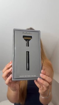 Kädet pitävät laatikkoon pakattua BaByliss Black Edition X-Blade -sähköparranajokonetta, jonka ajoterä ja kahva näkyvät leikkausten kautta.