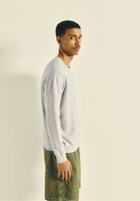 Pull léger gris clair avec des poignets côtelés, présentant une coupe décontractée. Associé à un short vert olive, mettant en valeur un look décontracté et superposé.