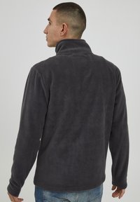 Mørkegrå fleece-pullover med høy krage, glatt tekstur, lange ermer og rett kant, sett bakfra.