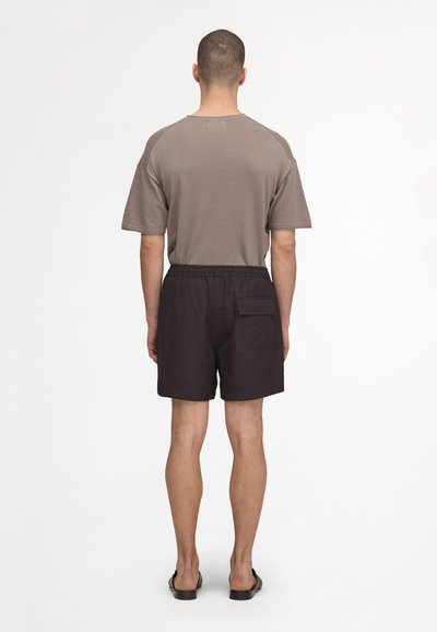 Homme debout, de dos, portant une chemise taupe à manches courtes, un short noir avec une poche arrière et des sandales noires sur fond blanc.