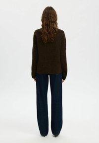 Donna con capelli lunghi e ricci che indossa un maglione morbido marrone scuro e jeans a gamba larga blu scuro, in piedi davanti a uno sfondo bianco semplice.