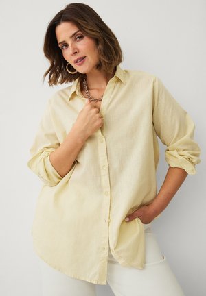 Vrouw met schouderlang bruin haar die een lichtgele blouse met knopen en een witte broek draagt, poseert met één hand in de zak en de andere bij de kraag.