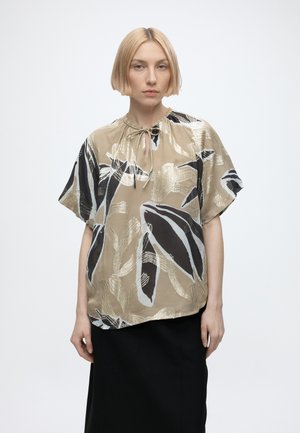 Mujer con cabello rubio corto que lleva una blusa suelta beige con un patrón abstracto de hojas en blanco y negro y una falda negra, de pie sobre un fondo liso.