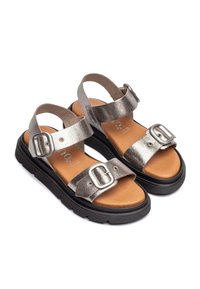 Sandales argentées métalliques avec des lanières ajustables, de grandes boucles, une semelle intérieure rembourrée marron et une épaisse semelle en caoutchouc noir.