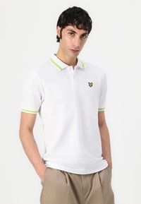 Polo shirt bianco a maniche corte, con colletto a righe verdi, due bottoni e un piccolo logo giallo ricamato sul petto.