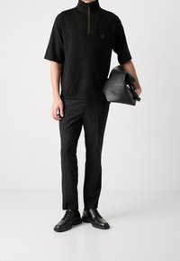 Černý krátký rukáv svetr s zipem a malým logem, zkombinovaný s černými elegantními kalhotami a lesklými černými šněrovacími botami, držící černou tašku.