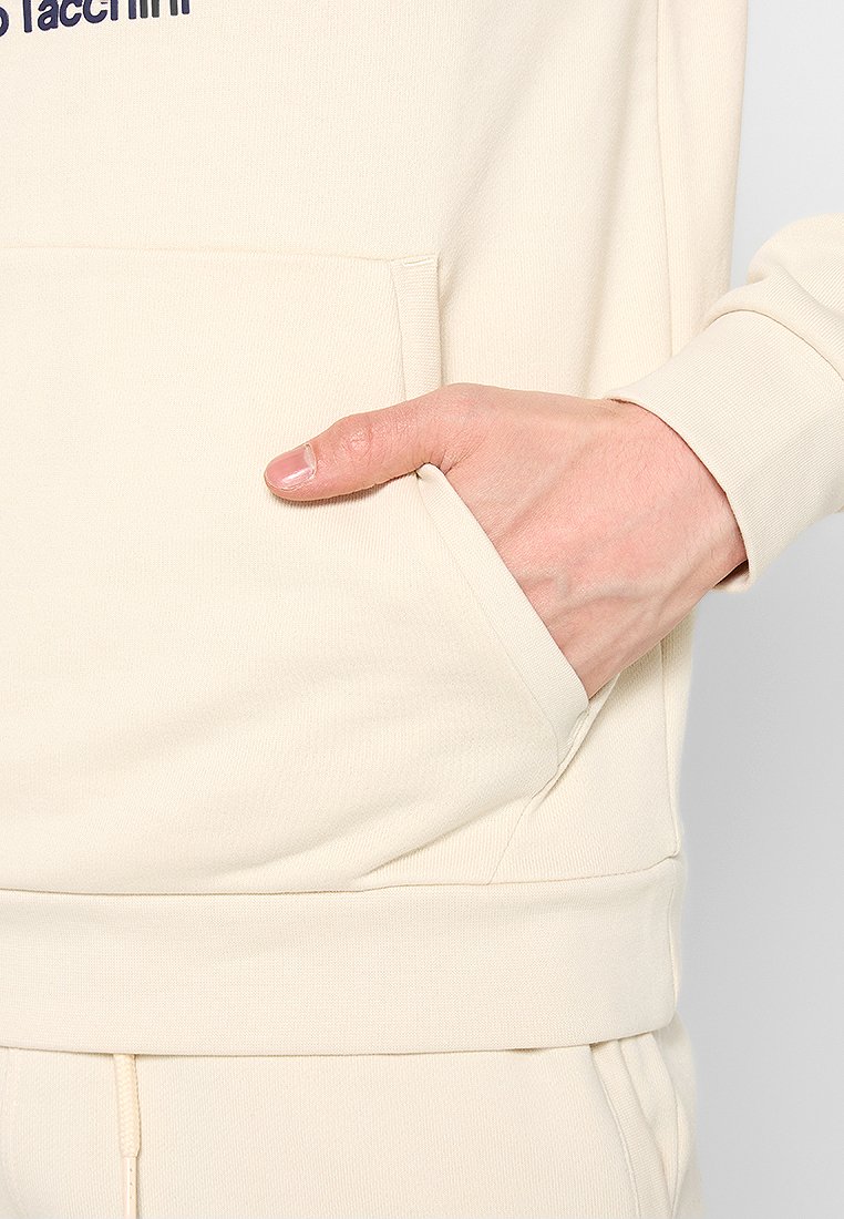 Sweatshirt couleur crème avec une poche avant. Détail en texte bleu foncé. Présente une coupe décontractée et des poignets côtelés. Tissu doux et texturé.