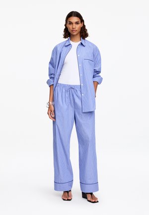 Femme portant une chemise ample rayée bleu et blanc et un pantalon large assorti, un haut blanc, des sandales à talons noires et un bracelet en argent.