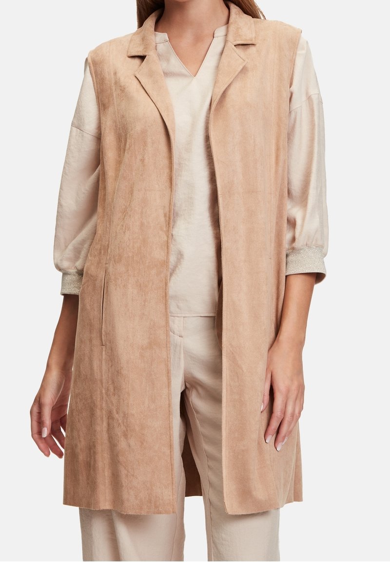 Beżowa zamszowa kamizelka bez rękawów z kołnierzem z klapami, przednimi kieszeniami i miękką fakturą. Noszona na neutralnym, dopasowanym stroju.