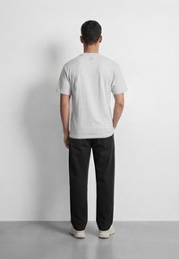 Άνδρας που στέκεται με το πρόσωπο προς έναν λευκό τοίχο, φορώντας ένα απλό ανοιχτό γκρι μπλουζάκι, μαύρο παντελόνι και λευκά αθλητικά παπούτσια, με τα χέρια χαλαρά στα πλευρά.