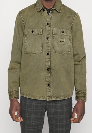 Mężczyzna ubrany w oliwkową koszulę na guziki z dwoma kieszeniami na piersi, z których jedna oznaczona jest napisem „BOSS”, oraz szare spodnie w kratę.