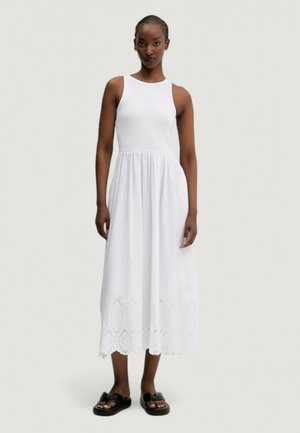 Abito bianco senza maniche con un corpino aderente e una gonna fluida, caratterizzato da ritagli ad occhiello all'orlo. La modella indossa sandali neri flats.