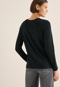 Dunkelgrüner Pullover mit langen Ärmeln aus weichem, strukturiertem Strick. Rundhalsausschnitt und figurbetonte Passform. Kombiniert mit hellgrauen, leicht verwaschenen Jeans.