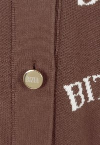 Bruin gebreid sweater met witte tekst en een metalen knop waarop "BIZUU" is gegraveerd. De stof heeft een fijne, gladde textuur.