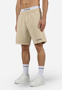 Pantalones cortos de algodón beige con cintura elástica, con bolsillos laterales y un logo negro "PEGADOR" en la pierna izquierda. Llevados con zapatillas blancas.